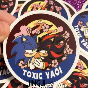 May include: Round sticker featuring blue and black cartoon characters with the text "TOXIC YAOI". The sticker has a dark red border with pink cherry blossoms and a yellow background. The sticker is held by a hand.
