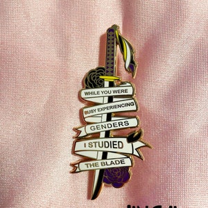 Puede incluir: Pin de esmalte dorado con una espada y una pancarta que dice "While you were busy experiencing genders I studied the blade". El pin está sobre un fondo rosa.
