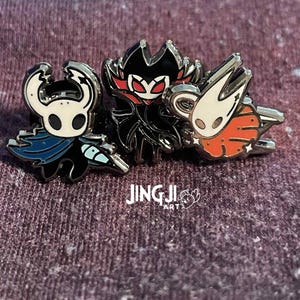 Puede incluir: Tres pines esmaltados con personajes del videojuego Hollow Knight. Los pines representan al Caballero, Grimm y un personaje con cuerpo naranja. Los pines tienen detalles en negro, blanco, azul y rojo. El texto "JING JI ART" es visible.