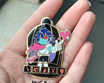 May include: A gold-colored enamel pin featuring a cartoon character with blue hair, a pink scarf, and a heart-shaped object. The pin has a black cage design and the words "ACT", "X", and a shield symbol. The pin is from "JINGJI ART".