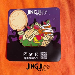 Puede incluir: Un colorido pin de esmalte con una tortuga y una magdalena con temática de Halloween. El pin tiene un fondo púrpura con una luna llena y siluetas. El pin tiene el texto "JingJi Art" e iconos de redes sociales.