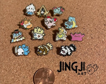 May include: A collection of 14 enamel pins featuring various cartoon characters. The pins are gold-toned and have a variety of colors, including blue, pink, yellow, and green. The pins are arranged on a corkboard surface with a penny for scale. The text "JINGJI ART" is visible in the lower right corner of the image.