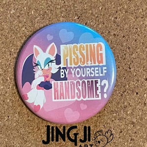 May include: A pink and blue button featuring a cartoon image of a white bat with pink ears and a pink nose. The bat has green eyes and is wearing a black choker. The text on the button reads "PISSING BY YOURSELF HANDSOME?" with a pink heart design around the text.