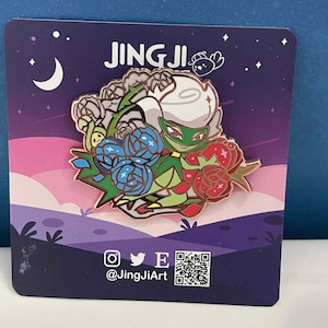 May include: A colorful enamel pin featuring a floral design with a character. The pin has a white, green, and red character surrounded by blue and red roses. The pin is on a dark purple card with the text "JING JI" and social media handles.