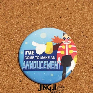 Könnte beinhalten: Ein runder Button mit blauem Hintergrund und dem Text "I'VE COME TO MAKE AN ANNOUNCEMENT". Er zeigt eine Zeichentrickfigur mit roten Haaren, Sonnenbrille und einer rot-weiß gestreiften Jacke. Der Button liegt auf einer braunen Oberfläche.