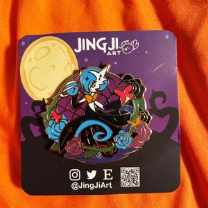 May include: A decorative enamel pin with a character design set against a full moon backdrop. The pin features a black, blue, and purple colour scheme with floral and butterfly details. The pin is on a black card with the text "JingJiArt".