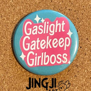 Peut inclure: Un bouton bleu et rose avec le texte "Gaslight Gatekeep Girlboss" en lettres blanches. Le bouton a un design scintillant.