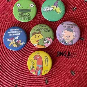 Pode incluir: Seis botões redondos coloridos com diferentes personagens de desenhos animados e texto. Os botões apresentam um sapo verde, uma criatura verde com um chapéu, um personagem amarelo com um amigo rosa, um personagem azul com um amigo vermelho, um personagem vermelho com uma gravata amarela e um homem com cabelo branco. O texto nos botões diz: "Hello I'm Mr Frog I Love You", "I Don't Do My Iconic Dance For Nothing", "I Think This Guy Is A Lost Cause", "That's A Little Version Of Me I Want You To Have It", e "JINGJI ART".