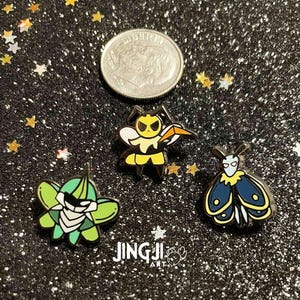 Op de afbeelding: Drie emaille pins met insectenontwerpen. De pins bevatten een groene en witte kever, een geelzwarte bij met een zwaard en een blauwgele mot. Naast de pins ligt een munt. De achtergrond is zwart met stervormige glitter.