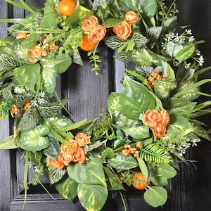May include: A vibrant wreath featuring lush green leaves with yellow accents, orange roses, and small orange fruit. The wreath is displayed on a black door, creating a striking contrast. A decorative piece for the home.