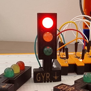 May include: A red, orange, and green traffic light with the red light illuminated. The traffic light is mounted on a black base with the word "GYR +" written on it. The traffic light is connected to a breadboard with wires.