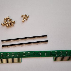 May include: A green circuit board with gold connectors and two black pin strips. There are also a pile of small gold colored metal pieces.