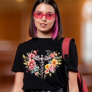 May include: A black t-shirt with a floral design featuring a medical caduceus symbol. The flowers are pink, yellow, and blue. The person wearing the shirt has pink hair and is wearing pink glasses.