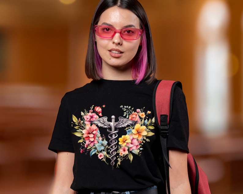 May include: A black t-shirt with a floral design featuring a medical caduceus symbol. The flowers are pink, yellow, and blue. The person wearing the shirt has pink hair and is wearing pink glasses.