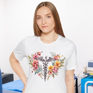 May include: White t-shirt with a floral design featuring a medical caduceus symbol in the center. The flowers are pink, yellow, and orange.