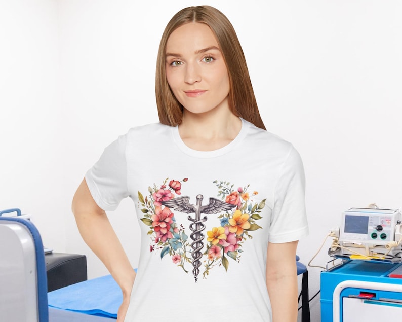 May include: White t-shirt with a floral design featuring a medical caduceus symbol in the center. The flowers are pink, yellow, and orange.