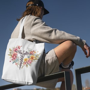 May include: A white tote bag with a floral design and a medical caduceus symbol. The bag is being held by a person wearing a grey sweatshirt and a black baseball cap.