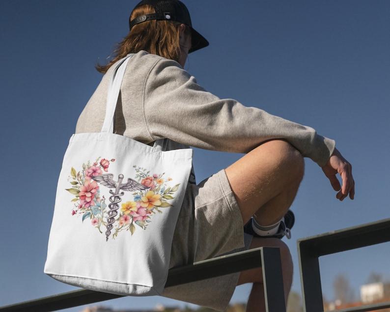 May include: A white tote bag with a floral design and a medical caduceus symbol. The bag is being held by a person wearing a grey sweatshirt and a black baseball cap.