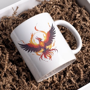 May include: White ceramic mug with a colorful phoenix rising from flames graphic. The mug is in a brown paper gift box.