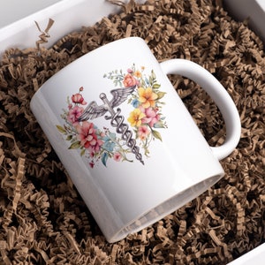 May include: White ceramic mug with a watercolor design of a medical caduceus surrounded by pink, yellow, and blue flowers.