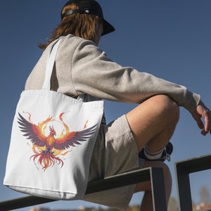 May include: A white tote bag with a colorful phoenix design. The phoenix is surrounded by flames and has a red, orange, and yellow color scheme.