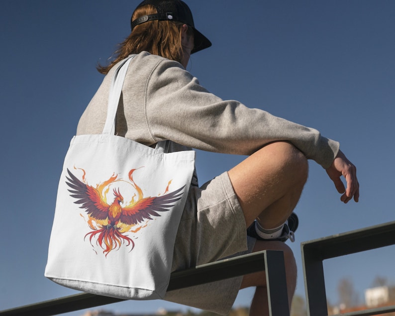 May include: A white tote bag with a colorful phoenix design. The phoenix is surrounded by flames and has a red, orange, and yellow color scheme.