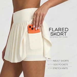 Op de afbeelding: Crèmekleurige uitlopende skort met ingebouwde shorts en zijzakken. De skort heeft een geplooid rokontwerp en een zak met een oranje telefoon. De tekst op de afbeelding luidt "FLARED SKORT SEWING PATTERN".