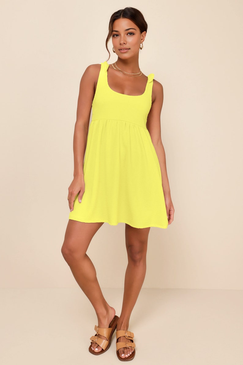 May include: A bright yellow sleeveless mini dress with a scoop neck and a tie detail at the shoulders. The dress has a slightly gathered waist and a flowy skirt.