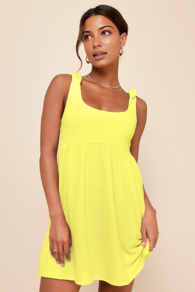 May include: A bright yellow sleeveless mini dress with a scoop neck and tie straps. The dress has a slightly gathered skirt.