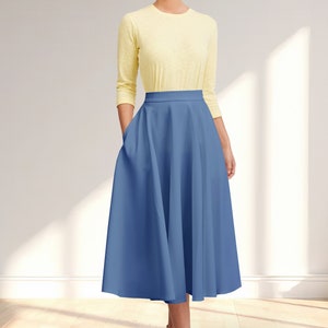 May include: A woman wearing a yellow long-sleeved shirt and a blue midi skirt with pockets. The skirt has a high waist and a full, flowing silhouette.