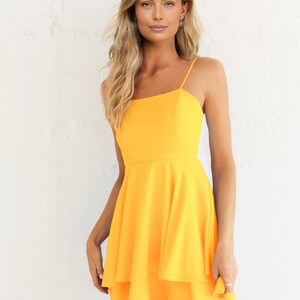 May include: A bright yellow dress with a square neckline and spaghetti straps. The dress features a fitted bodice and a flared, layered skirt. The model is wearing the dress in front of a white wall.