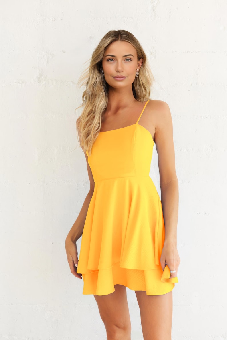 May include: A bright yellow dress with a square neckline and spaghetti straps. The dress features a fitted bodice and a flared, layered skirt. The model is wearing the dress in front of a white wall.