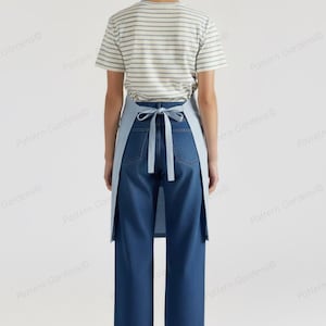 Puede incluir: Un delantal azul claro con una correa para el cuello y lazos en la cintura, usado sobre una camiseta a rayas y unos vaqueros azules. El delantal es de color liso y los vaqueros son de lavado oscuro. La persona lleva sandalias.