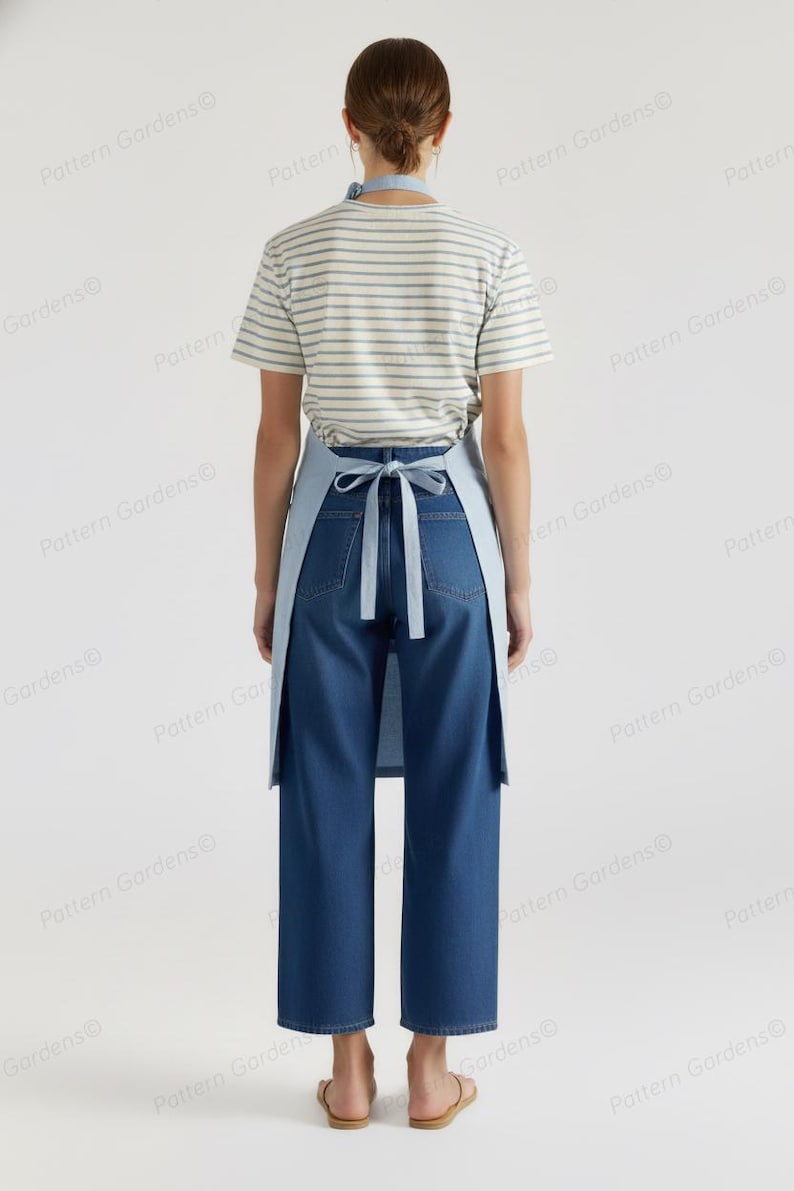 Puede incluir: Un delantal azul claro con una correa para el cuello y lazos en la cintura, usado sobre una camiseta a rayas y unos vaqueros azules. El delantal es de color liso y los vaqueros son de lavado oscuro. La persona lleva sandalias.