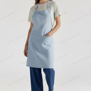 Puede incluir: Un delantal azul claro con un bolsillo delantero grande y correas ajustables. El delantal se usa sobre una camiseta a rayas y pantalones azul oscuro. La persona lleva sandalias doradas.