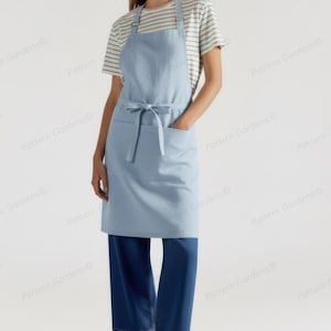 Puede incluir: Un delantal azul claro con un bolsillo delantero y un lazo en la cintura. El delantal se lleva sobre una camiseta a rayas y pantalones azules. El delantal est&aacute; hecho de una tela suave y duradera.
