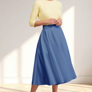 May include: A woman wearing a yellow 3/4 sleeve shirt and a blue midi skirt. She is standing on a wooden floor with natural light coming in from a window.