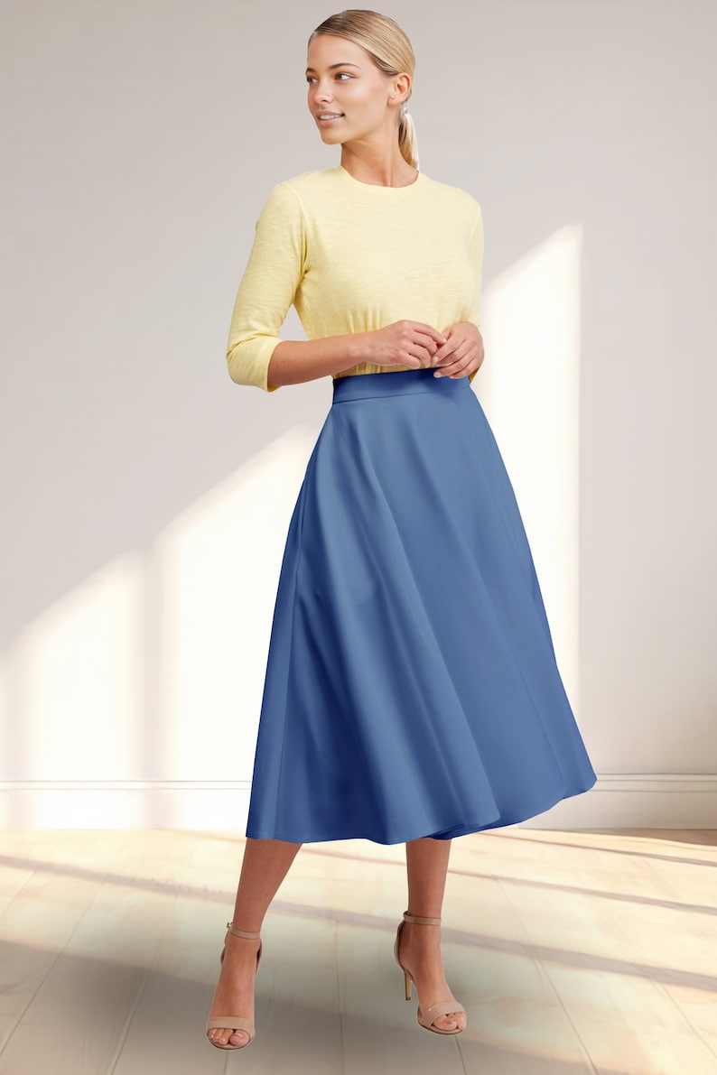 May include: A woman wearing a yellow 3/4 sleeve shirt and a blue midi skirt. She is standing on a wooden floor with natural light coming in from a window.