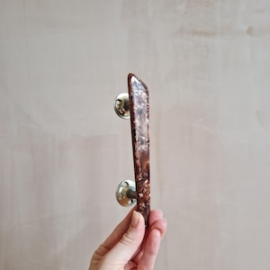 May include: A brown and gold cabinet pull with a rectangular handle. The handle has a marbled appearance with gold flecks and is attached to two circular, brass-coloured mounting plates.