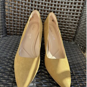 May include: A pair of mustard-yellow Clarks pumps with a textured pattern. The shoes have pointed toes and a low heel. The Clarks logo is visible inside the shoe.