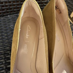 May include: Close-up of two mustard-colored Clarks brand flats. The interior of the shoe is a light tan color with the brand name and "cushion soft" printed on the insole. The exterior has a textured pattern.