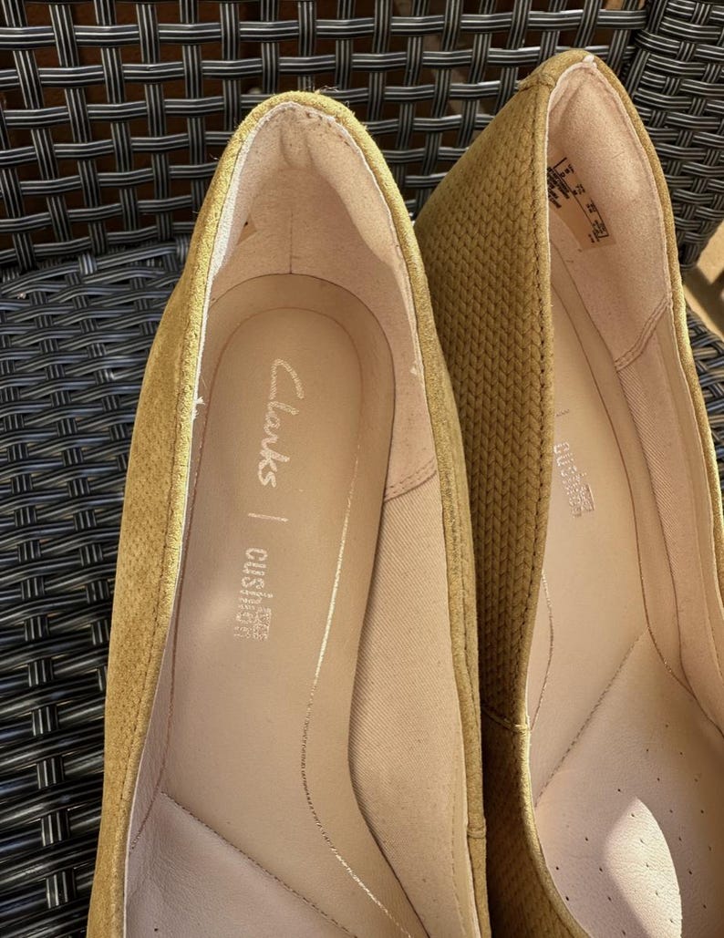 May include: Close-up of two mustard-colored Clarks brand flats. The interior of the shoe is a light tan color with the brand name and "cushion soft" printed on the insole. The exterior has a textured pattern.