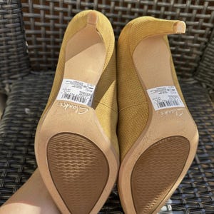 May include: A pair of mustard yellow high-heeled shoes with pointed toes. The shoes have a textured fabric upper and a slender heel. The soles are a light brown color. The brand name "Clarks" is visible on the sole.