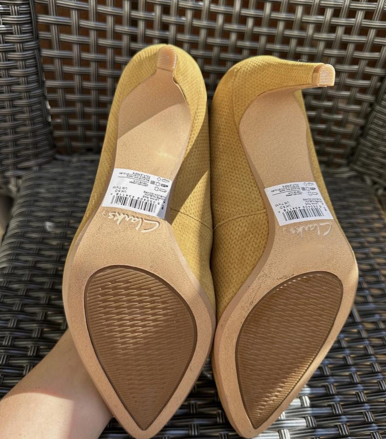 May include: A pair of mustard yellow high-heeled shoes with pointed toes. The shoes have a textured fabric upper and a slender heel. The soles are a light brown color. The brand name "Clarks" is visible on the sole.