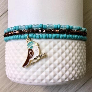 May include: A set of three turquoise and brown beaded bracelets with a small bird charm. The bird charm is wearing a blue hat and holding a small branch.