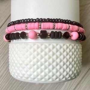 May include: Three layered bracelets with pink, black, and dark purple beads. The bracelets are stacked on top of each other.