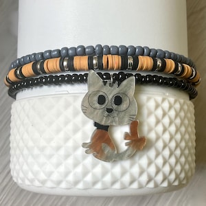 May include: Beaded bracelets with a cat charm. The bracelets have grey, orange, black, and silver beads. A silver cat charm with orange and brown details hangs from the bracelets. The bracelets are displayed on a white, textured surface.