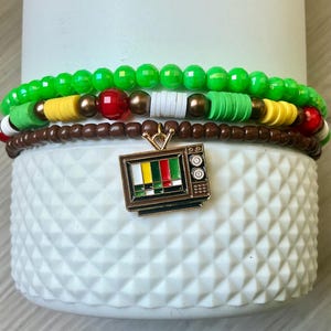 Puede incluir: Conjunto de pulseras de cuentas en tonos verdes, marrones, amarillos, rojos y blancos. Las pulseras tienen una variedad de formas y tamaños de cuentas. Un dije dorado en forma de televisor vintage está unido a la pulsera inferior.