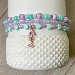 Könnte beinhalten: Ein weißer Becher mit drei Lagen violetter und grüner Perlenarmbänder. Ein violetter und goldener Fisch-Anhänger hängt an der Unterseite der Armbänder.