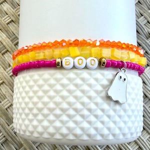 Könnte beinhalten: Ein Stapel aus drei Perlenarmbändern mit einem Strang aus orangefarbenen facettierten Perlen, einem Strang aus gelben quadratischen Perlen und einem Strang aus pinkfarbenen Saatperlen. Das pinke Armband enthält weiße Buchstabenperlen mit der Aufschrift BOO und einen silbernen Geisteranhänger.